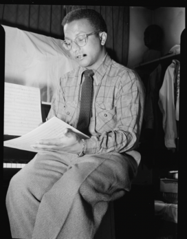 Billy Strayhorn is a young-looking man in shirtsleeves and necktie. He sits near a piano and seems to be looking through sheet music.