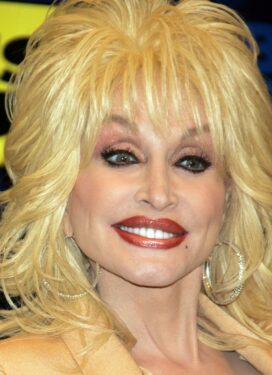 Photo of Dolly Parton in hoop earrings and a big ol' head of platinum blonde hair, smiling. We can just see the top of her gold jacket